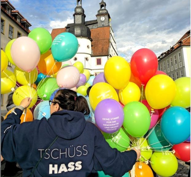 Eine Person mit bunten Luftballons trägt einen Pullover mit der Aufschrift "TSCHÜSS HASS"