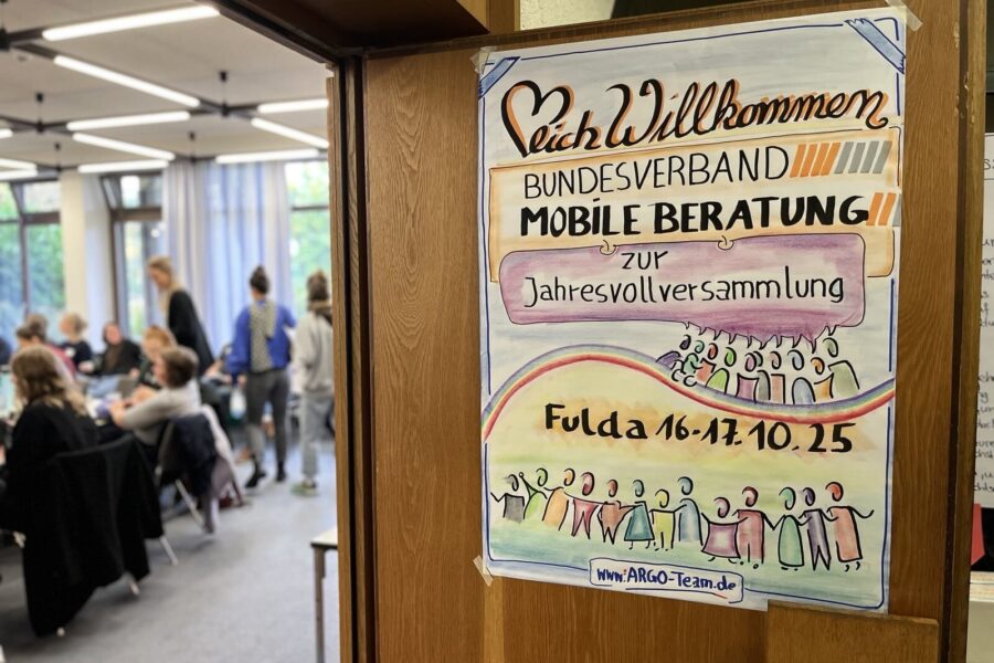 Rechts ein buntes Poster, das an einer Tür hängt und auf dem steht: "Herzlich Willkommen zur Jahresvollversammlung des Bundesverbands Mobile Beratung." Fulda 16.-17.10.25. Links im Hintergrund sind verschwommen Personen zu sehen.